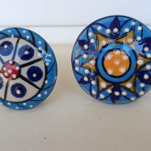 Peut inclure: Deux boutons de placard ronds en c&eacute;ramique avec des motifs peints &agrave; la main. Le bouton de gauche pr&eacute;sente un centre cr&egrave;me avec des cercles bleus et un point rouge, tandis que celui de droite affiche un centre jaune entour&eacute; de p&eacute;tales bleus.