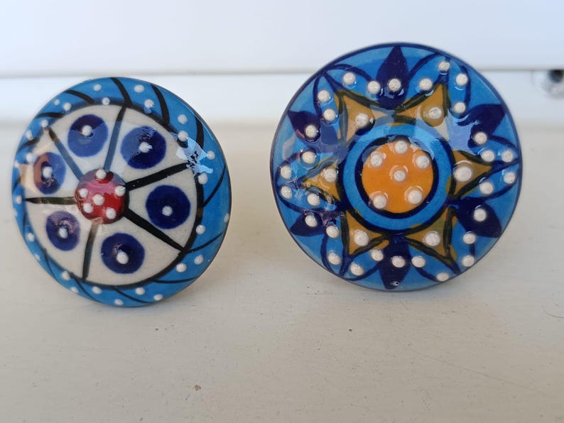 Peut inclure: Deux boutons de placard ronds en c&eacute;ramique avec des motifs peints &agrave; la main. Le bouton de gauche pr&eacute;sente un centre cr&egrave;me avec des cercles bleus et un point rouge, tandis que celui de droite affiche un centre jaune entour&eacute; de p&eacute;tales bleus.