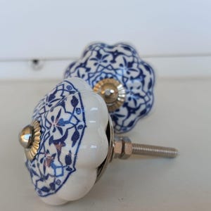 Peut inclure: Deux boutons de placard ronds en c&eacute;ramique avec un bord festonn&eacute;, une base blanche et un motif floral bleu d&eacute;taill&eacute;. Chaque bouton poss&egrave;de un accent central en m&eacute;tal argent&eacute; et une tige filet&eacute;e m&eacute;tallique &agrave; l'arri&egrave;re pour la fixation.
