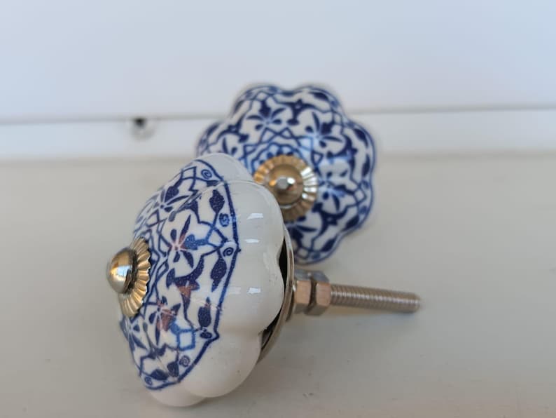 Peut inclure: Deux boutons de placard ronds en c&eacute;ramique avec un bord festonn&eacute;, une base blanche et un motif floral bleu d&eacute;taill&eacute;. Chaque bouton poss&egrave;de un accent central en m&eacute;tal argent&eacute; et une tige filet&eacute;e m&eacute;tallique &agrave; l'arri&egrave;re pour la fixation.