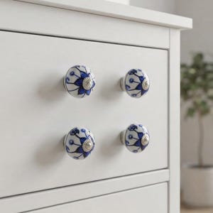 May include: Four round ceramic drawer knobs attached to a white dresser. Each knob features a white base with a hand-painted blue floral pattern and a silver-toned metal center accent. The knobs are arranged in two rows of two on the drawer front.