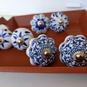 Peut inclure: Un ensemble de six boutons de placard en c&eacute;ramique pr&eacute;sentant des surfaces blanches avec des motifs peints en bleu. Les boutons varient en forme, incluant des designs ronds et festonn&eacute;s, chacun dot&eacute; d'une pi&egrave;ce m&eacute;tallique argent&eacute;e.