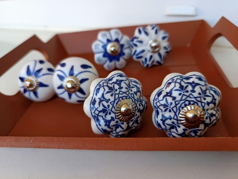 Peut inclure: Un ensemble de six boutons de placard en c&eacute;ramique pr&eacute;sentant des surfaces blanches avec des motifs peints en bleu. Les boutons varient en forme, incluant des designs ronds et festonn&eacute;s, chacun dot&eacute; d'une pi&egrave;ce m&eacute;tallique argent&eacute;e.