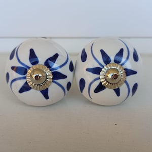 Peut inclure: Deux boutons de placard ronds en c&eacute;ramique avec une base couleur cr&egrave;me et un motif floral bleu fonc&eacute; peint &agrave; la main. Chaque bouton poss&egrave;de un capuchon central en m&eacute;tal dor&eacute; brillant avec un bord festonn&eacute;, ajoutant une touche d&eacute;corative.