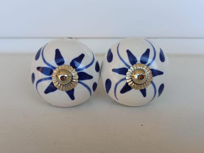 Peut inclure: Deux boutons de placard ronds en c&eacute;ramique avec une base couleur cr&egrave;me et un motif floral bleu fonc&eacute; peint &agrave; la main. Chaque bouton poss&egrave;de un capuchon central en m&eacute;tal dor&eacute; brillant avec un bord festonn&eacute;, ajoutant une touche d&eacute;corative.