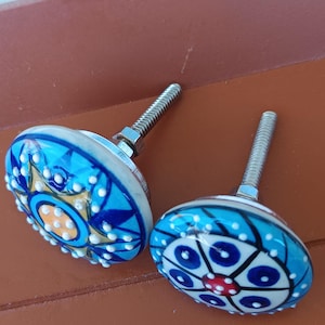 Peut inclure: Deux boutons de placard ronds en c&eacute;ramique avec vis et &eacute;crous en m&eacute;tal. Le bouton de gauche pr&eacute;sente un motif d'&eacute;toile bleu, jaune et blanc avec des points blancs en relief. Le bouton de droite affiche un motif floral circulaire bleu, blanc et rouge.