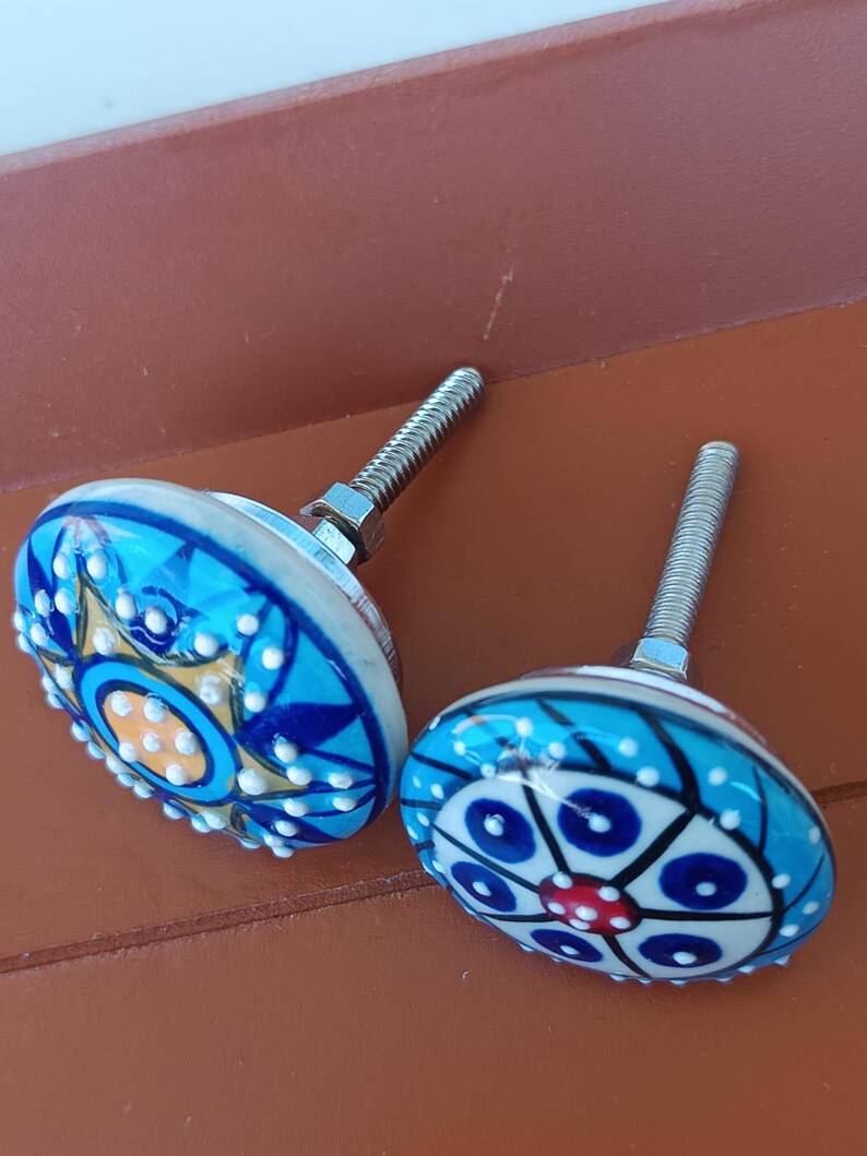 Peut inclure: Deux boutons de placard ronds en c&eacute;ramique avec vis et &eacute;crous en m&eacute;tal. Le bouton de gauche pr&eacute;sente un motif d'&eacute;toile bleu, jaune et blanc avec des points blancs en relief. Le bouton de droite affiche un motif floral circulaire bleu, blanc et rouge.
