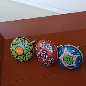 Peut inclure: Trois boutons de placard ronds en c&eacute;ramique avec des vis m&eacute;talliques. Le bouton de gauche est vert avec un centre jaune et des points orange. Le bouton central est rouge avec des motifs floraux blancs. Celui de droite est bleu et blanc.