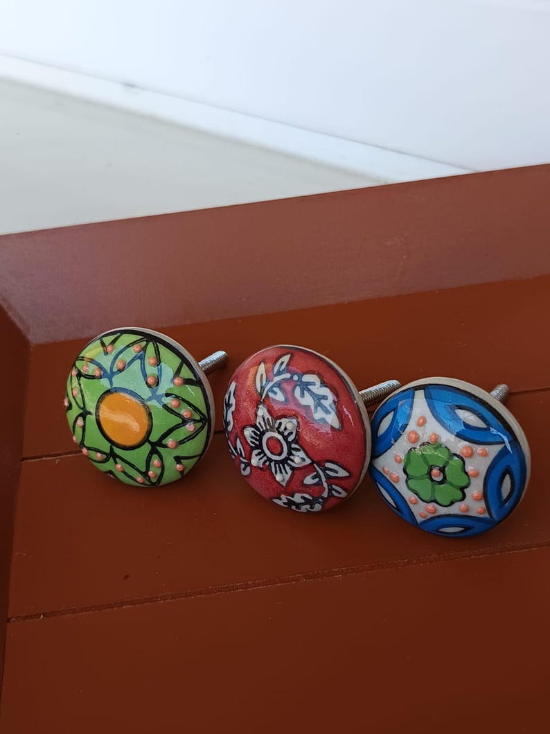 Peut inclure: Trois boutons de placard ronds en c&eacute;ramique avec des vis m&eacute;talliques. Le bouton de gauche est vert avec un centre jaune et des points orange. Le bouton central est rouge avec des motifs floraux blancs. Celui de droite est bleu et blanc.