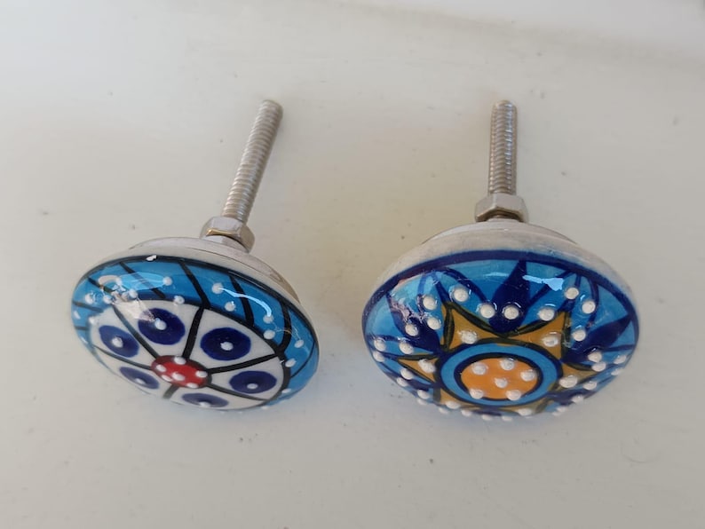 Peut inclure: Deux boutons de placard ronds en c&eacute;ramique avec des motifs peints &agrave; la main. Le bouton de gauche pr&eacute;sente un motif g&eacute;om&eacute;trique bleu, blanc et rouge. Le bouton de droite affiche un motif en &eacute;toile bleu, jaune et blanc. Inclut boulon et &eacute;crou.
