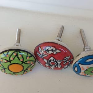 Peut inclure: Trois boutons de placard ronds en c&eacute;ramique avec des vis m&eacute;talliques. Le bouton de gauche est vert clair avec un cercle orange central et des d&eacute;tails noirs. Le bouton central est rouge avec des motifs floraux blancs. Celui de droite est blanc.