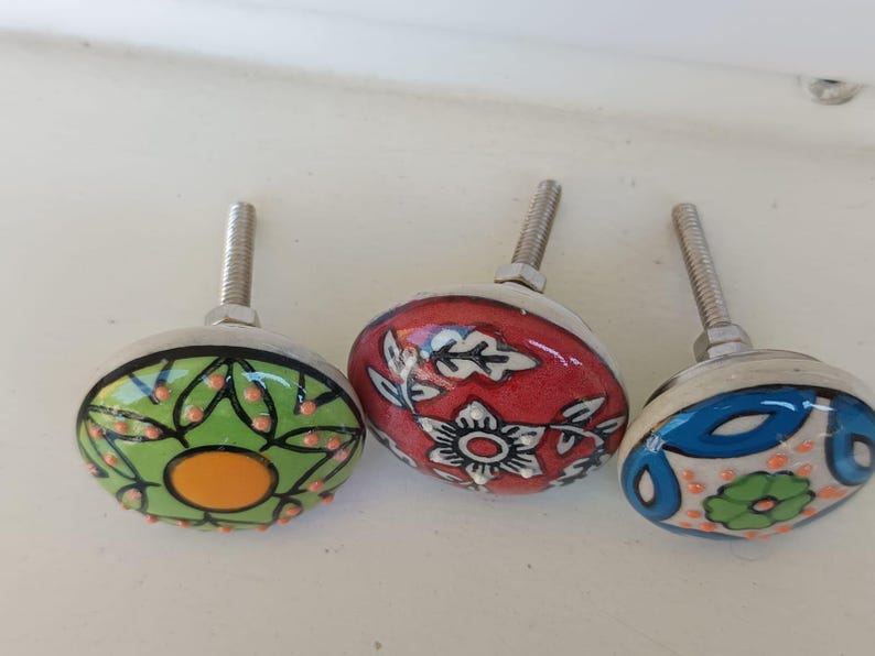 Peut inclure: Trois boutons de placard ronds en c&eacute;ramique avec des vis m&eacute;talliques. Le bouton de gauche est vert clair avec un cercle orange central et des d&eacute;tails noirs. Le bouton central est rouge avec des motifs floraux blancs. Celui de droite est blanc.