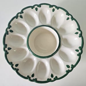 May include: A round white ceramic devilled egg platter featuring twelve oval indentations for eggs and a central circular bowl. The rim is decorated with a scalloped edge and a dark green painted border with small teardrop accents.