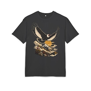 Op de afbeelding: Een antracietgrijs T-shirt met korte mouwen en een grafische print van een grote vogel met zwart-witte vleugels die boven besneeuwde bergtoppen, wolken en een gouden zon vliegt. Het ontwerp is gecentreerd op de borst van het kledingstuk.