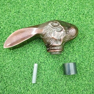 Puede incluir: Un tirador de mueble de tono cobre metálico con forma de cabeza de conejo y una oreja larga que se extiende hacia la izquierda. El tirador presenta texturas de pelaje detalladas y un ojo oscuro. Incluye un perno roscado plateado y un pequeño cilindro negro.