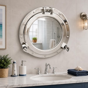 May include: A circular porthole mirror with a polished chrome finish, featuring a hinged top latch and two side locking handles. The frame is secured with decorative rivets around the perimeter, reflecting a bathroom interior with a sink.