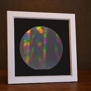May include: A circular silicon wafer featuring a grid of microchips with a holographic, iridescent finish displaying vibrant rainbow colours, encased in a square white frame with a black mat, presenting a technical and scientific aesthetic.