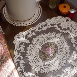 Peut inclure: Un napperon en dentelle de forme ovale couleur crème présentant un motif floral complexe avec des roses roses et des feuilles vertes brodées au centre. La dentelle possède un fond en maille délicat et une bordure festonnée.