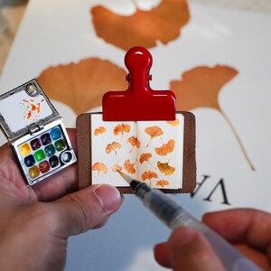 Può includere: Un taccuino per schizzi in miniatura con copertina in pelle marrone tenuto da una clip in metallo rosso brillante. Le pagine aperte presentano foglie di ginkgo arancioni dipinte a mano. Accanto c'è una minuscola tavolozza per acquerelli.