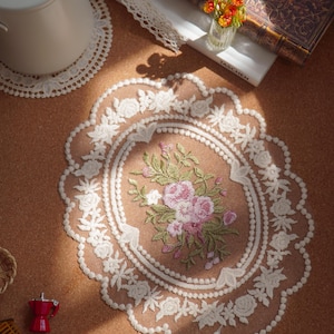 Könnte beinhalten: Eine ovale Spitzendeckchen mit einem komplizierten weißen Blumenrand und einem zentralen gestickten Motiv aus rosa und weißen Blumen mit grünen Blättern, platziert auf einer braunen Oberfläche mit anderen dekorativen Haushaltsgegenständen.