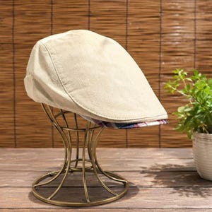 May include: A beige flat cap featuring a structured crown and a short, stiff brim. The underside of the brim displays a multi-colored plaid pattern in shades of red, blue, and white. The cap is displayed on a gold-toned wire stand.