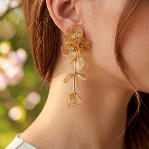 May include: A long gold-toned earring featuring a vertical arrangement of delicate, textured metal flower petals and leaves. The design includes multiple layered floral elements with small, clear, sparkling crystals set in the centers of the top blooms.