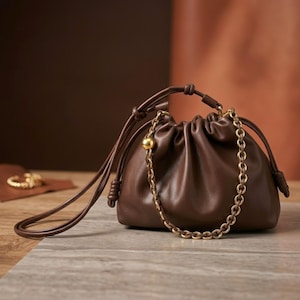 May include: A dark brown leather drawstring pouch bag featuring a gathered top closure, a long thin shoulder strap with knotted ends, and a decorative gold-tone metal chain handle accented with a single polished gold sphere bead on the side.