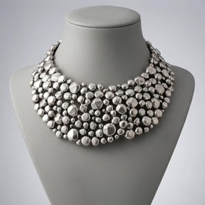 May include: A statement collar necklace featuring a dense arrangement of polished silver-toned metallic pebbles of varying sizes. The rounded, irregular shapes are clustered together to form a wide, textured bib design that sits around the neck.