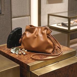 Può includere: Una borsa a sacchetto in pelle color cuoio con una silhouette morbida e arricciata, dotata di chiusura a coulisse con estremità annodate e una tracolla lunga e sottile. La borsa presenta una finitura liscia e opaca e una forma arrotondata.