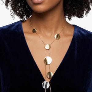 May include: A gold-toned lariat necklace featuring a series of polished circular discs of varying sizes suspended along thin metallic chains. The design includes multiple round elements arranged in a vertical drop pattern against a dark blue fabric.