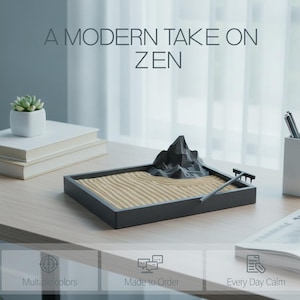 May include: A rectangular black tray containing light beige sand, a dark gray geometric mountain sculpture, and a small black rake. The text A MODERN TAKE ON ZEN is visible above the tray. The set includes icons for multiple colors, made to order, and every day calm.
