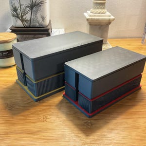 May include: Two rectangular storage boxes with dark grey lids and bodies. The boxes feature vertical ribbed detailing on the lower sections. One box has a gold-coloured base accent, while the other box features a red-coloured base accent.