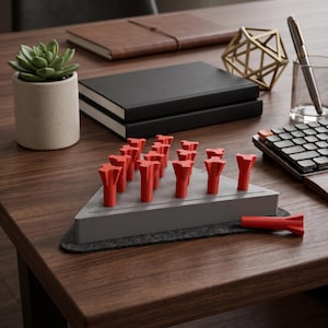 May include: A triangular gray game board featuring a series of holes with several bright red T-shaped pegs inserted into the board. One additional red T-shaped peg lies horizontally on the dark gray felt base beneath the game board structure.