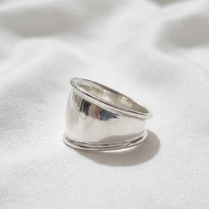 May include: A wide, polished silver ring featuring a smooth, concave band with raised, rounded edges at the top and bottom. The metal surface is highly reflective, showing subtle highlights and shadows across its curved, solid construction.