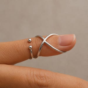 Könnte beinhalten: Ein silberfarbener Ring mit einem überkreuzten Design, das sich um den Finger legt. Das Band besteht aus zwei dünnen, glatten Metalldrähten, die sich zu einem X kreuzen und auf einer Seite in zwei kleinen, polierten Kugeln enden.