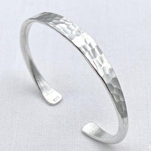 May include: A silver cuff bracelet with a hammered, textured finish on the outer surface. The bracelet features an open design with rounded ends, and the interior of one end is clearly stamped with the numerical text 925, indicating sterling silver.