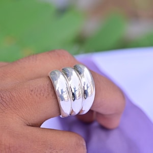 May include: A polished silver-toned ring featuring three thick, smooth, rounded bands fused together into a single wide piece. The metal surface is highly reflective, showing light highlights and a curved, continuous shape across the finger.