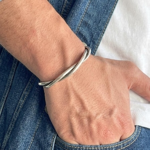 Può includere: Un bracciale a fascia in metallo color argento con una finitura liscia e lucida, caratterizzato da un design intrecciato e sovrapposto che crea un effetto ad anello continuo attorno al polso, con una superficie riflettente e bordi arrotondati.