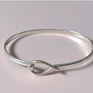 May include: A polished silver-tone bangle bracelet featuring a smooth, continuous band that loops into a teardrop-shaped hook closure at the front. The metal surface is reflective and uniform, creating a clean, circular silhouette for the wrist.