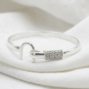 May include: A polished silver-tone bangle bracelet featuring a hook-and-eye style clasp mechanism. The design includes a smooth, rounded band that transitions into a textured, twisted wire detail near the hook closure, creating a clean look.