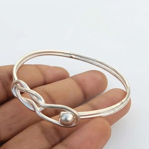 Könnte beinhalten: Ein polierter Armreif aus Silber mit einem durchgehenden, glatten Metallband, das an der Vorderseite zu einem Achterknoten geformt ist. Der Armreif hat eine reflektierende, metallische Oberfläche und ein abgerundetes, schmales Profil.