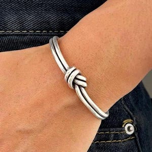 May include: A silver-toned metal cuff bracelet featuring a double-strand design that converges into a central knot detail. The bracelet has a smooth, polished finish and a rounded profile, creating a continuous loop around the wrist area.