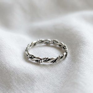 May include: A silver ring featuring a twisted rope design with an oxidised finish that highlights the intricate texture of the metal strands. The band has a continuous, uniform pattern that wraps around the entire circumference of the circular ring.