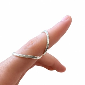 Könnte beinhalten: Ein silberfarbener Metallring mit einer texturierten, gehämmerten Oberfläche. Das Band hat ein unregelmäßiges, facettiertes Aussehen, das durch den Hammervorgang entsteht und das Licht auf der metallischen Oberfläche reflektiert. Der Ring wird am Finger getragen.