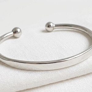 Könnte beinhalten: Ein poliertes silberfarbenes Armband in Manschettenform mit einem glatten, offenen Banddesign und einer kleinen, abgerundeten Kugel an jedem Ende. Die Metalloberfläche ist reflektierend und gleichmäßig und bildet eine klare, runde Form.
