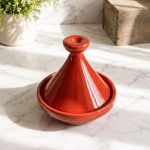 Peut inclure: Un tajine en céramique de couleur terre cuite brillante, doté d'une large base circulaire et d'un couvercle conique haut surmonté d'une poignée ronde. Ce récipient de cuisson présente une finition lisse et réfléchissante ainsi qu'une forme conique.