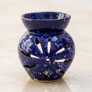 May include: A deep blue ceramic oil burner with a glossy, speckled finish. The spherical body features a cut-out flower design with eight petals, and a wide, bowl-shaped top section for holding wax melts or essential oils and water for diffusion.