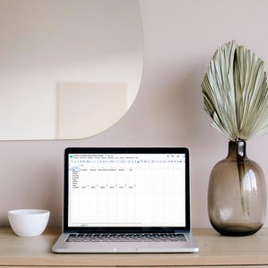 Op de afbeelding: Een zilveren laptop met een Etsy Seller Performance Tracker-spreadsheet op het scherm, naast een kleine witte kom en een bruine glazen vaas met een gedroogd palmblad, geplaatst op een lichthouten ondergrond.