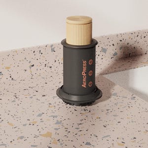 May include: A cylindrical coffee maker with a matte black body and a light tan plunger. The word AEROPRESS is printed vertically in orange on the side, accompanied by the numbers 1, 2, and 3, each enclosed in an orange circle.