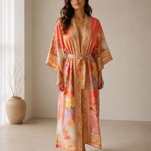 Puede incluir: Una bata larga y fluida con un estampado floral vibrante en tonos coral, naranja y morado suave. La prenda incluye mangas anchas, un cinturón de tela a juego en la cintura y un borde decorativo a lo largo de la abertura frontal y el dobladillo.